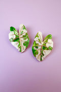 Two decorative pins shaped like monstera leaves, inspired by hand-crafted jewelry, feature a mix of green and white colors with gold vein detailing, and are placed on a light purple background.