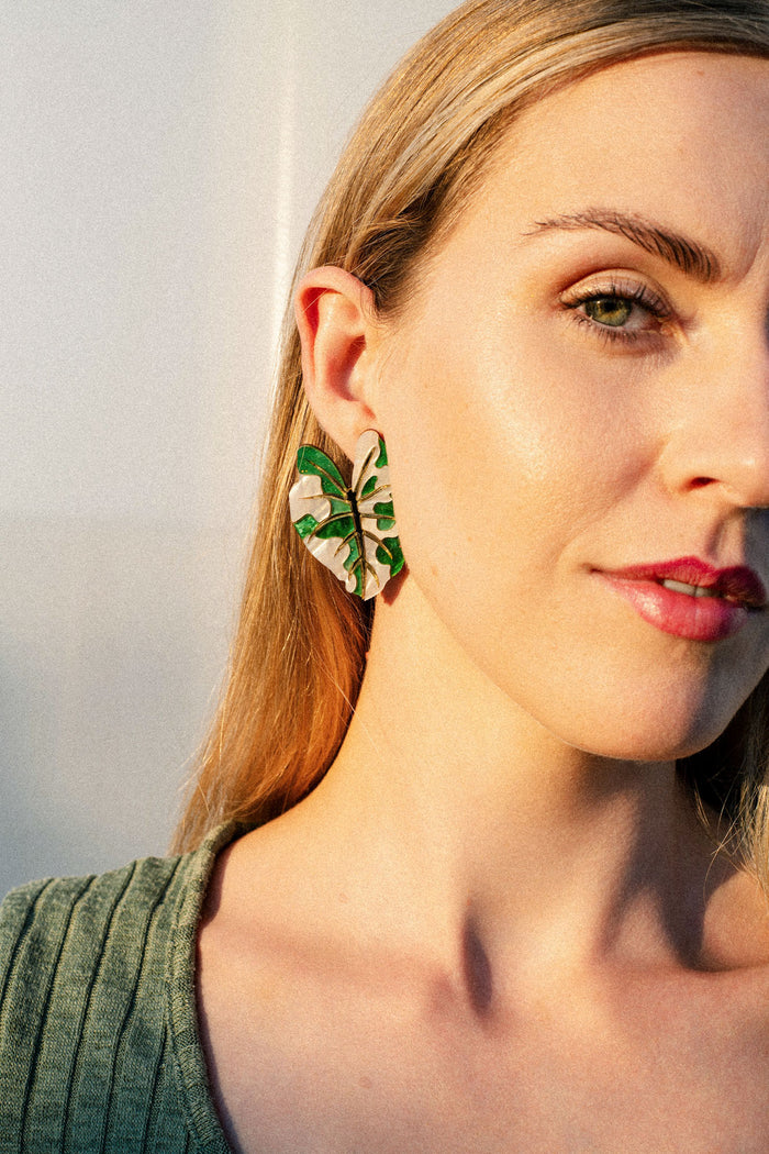 A woman with blonde hair wears a green ribbed top and a large Alocasia Earrings in a green and beige pattern. She is facing the camera in soft, natural light, showcasing the beauty of this hand-crafted jewelry.