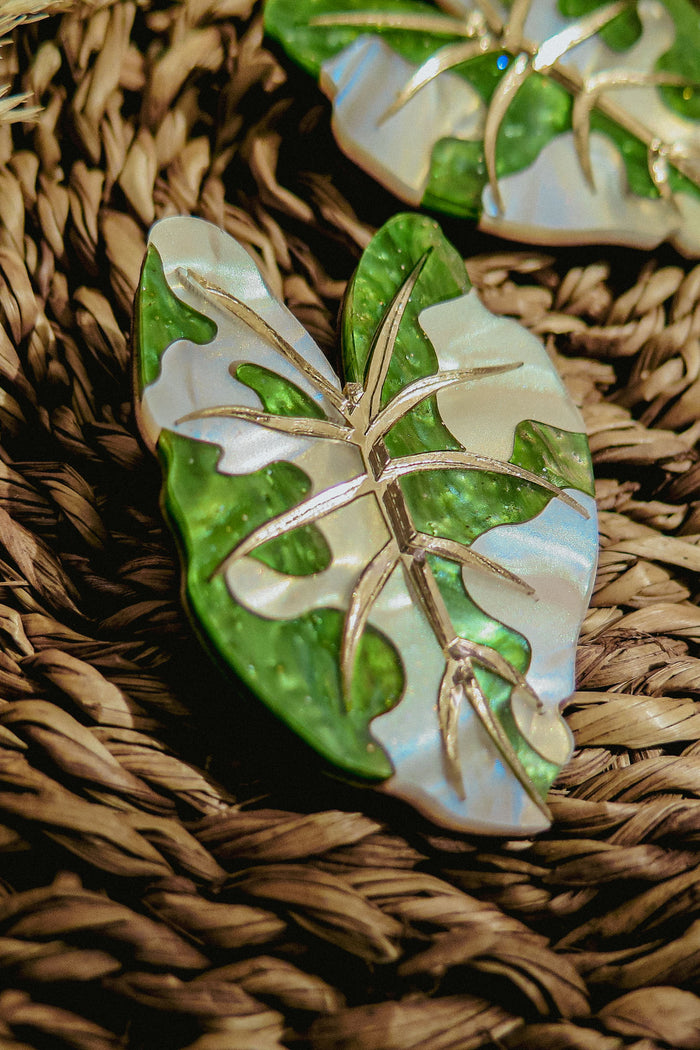 A close-up of Alocasia Earrings, shaped like a green and white leaf with metallic veins, resting on a woven, textured surface—perfect for those who love hand-crafted jewelry.