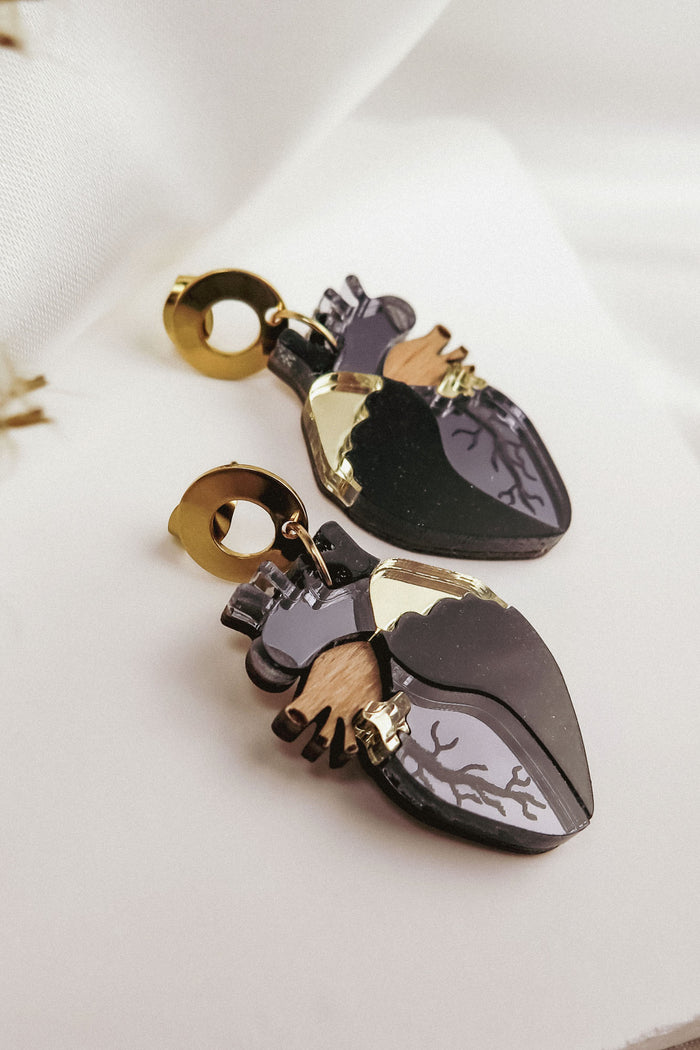 A pair of Anatomical Heart Earrings with gold accents and black, grey, and wood-toned details adds a touch of gothic flair, displayed on a white fabric background—perfect for lovers of unique handcrafted accessories.
