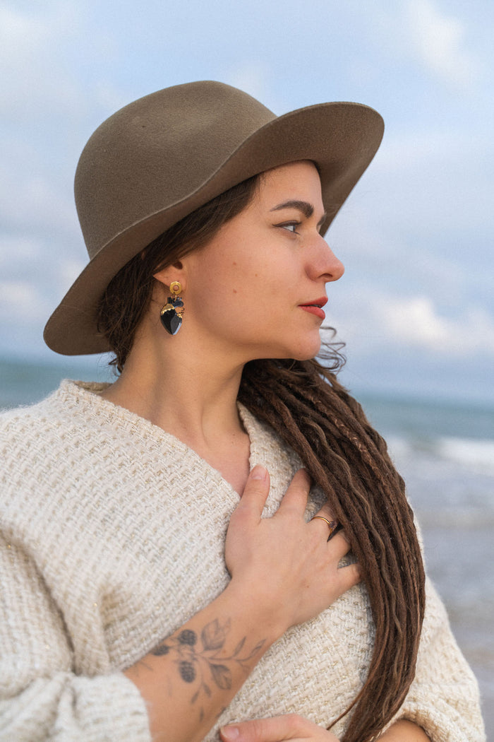 A woman with long brown dreadlocks, wearing a wide-brimmed hat, cream sweater, and handcrafted Anatomical Heart Earrings, stands by the sea with one hand on her chest. She has a tattoo on her forearm and looks serene.
