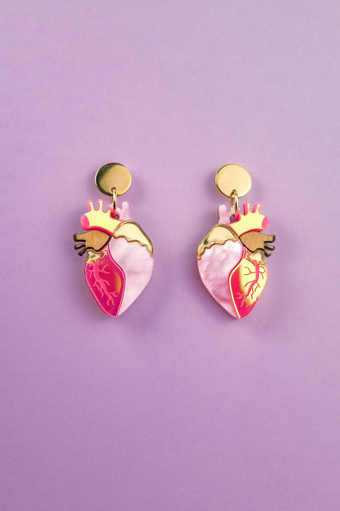 A pair of bold style Anatomical Heart Earrings crafted from holographic acrylic, with gold-toned tops and pink and red details, displayed on a pastel purple background.