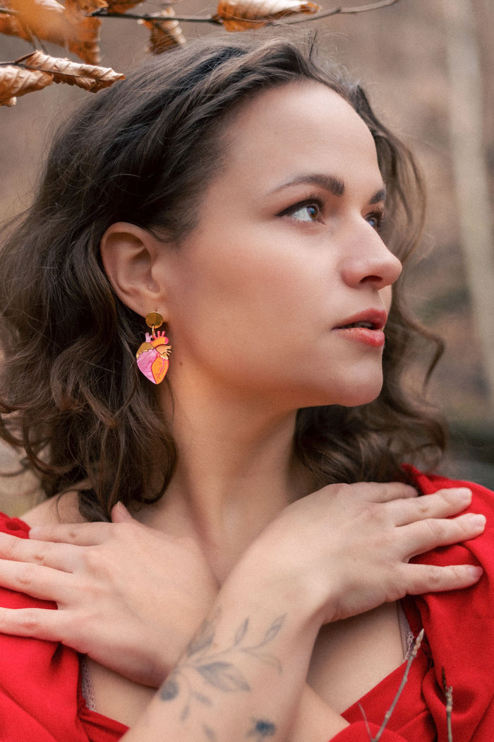 A woman with wavy brown hair and a tattooed forearm crosses her hands over her chest. She wears a red dress, bold style pink statement earrings, and stands outdoors among brown autumn leaves, looking to the side.