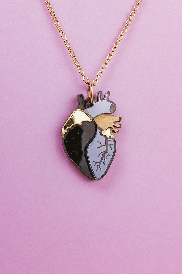 A gold chain anatomical heart necklace with a pendant featuring black, gray, and gold sections is displayed against a pink background—a truly unique jewelry piece.