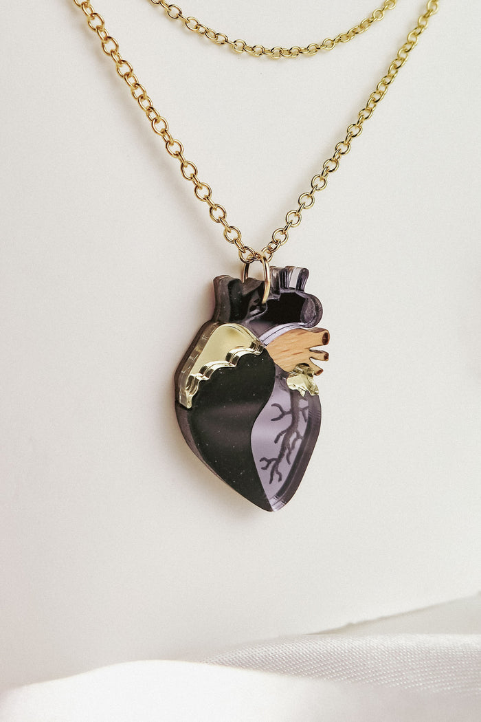 A gold chain necklace with a pendant shaped like an anatomical heart, featuring black, gold, and wood-toned sections etched with vein-like details. This handcrafted jewelry piece rests against a white background.