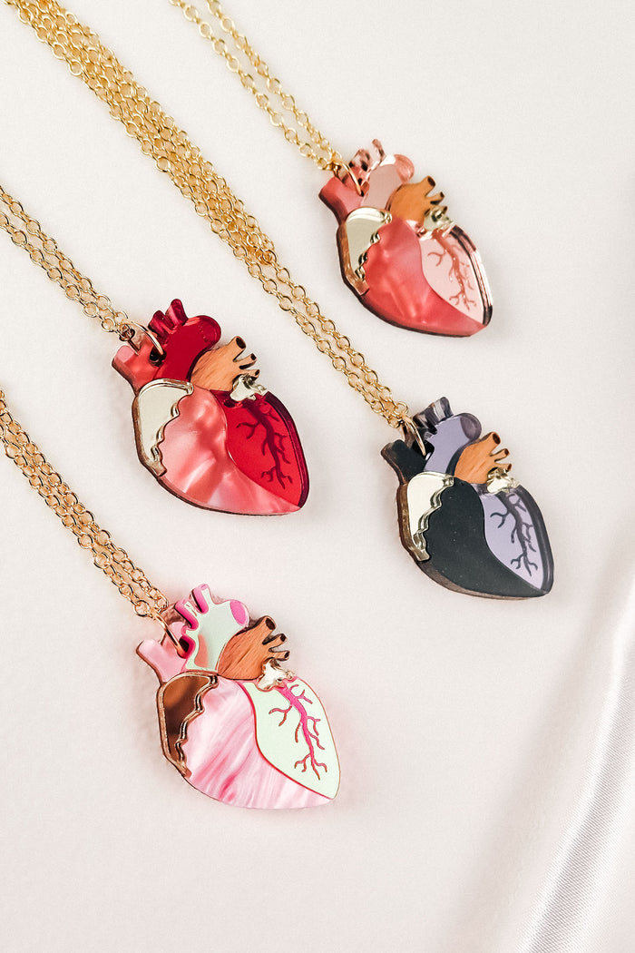 Four handcrafted jewelry necklaces with gold chains, each featuring an anatomical heart necklace pendant in dark red, pink, black and purple, or pink and mint green—making each one a unique jewelry piece displayed on a light background.