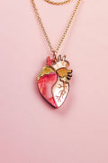 A handcrafted gold chain necklace with a pendant shaped like an anatomical human heart, featuring pink, gold, and cream accents—this unique jewelry piece is beautifully displayed on a pale pink background.