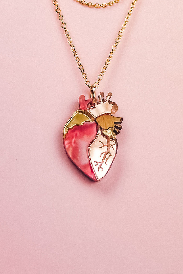 A handcrafted gold chain necklace with a pendant shaped like an anatomical human heart, featuring pink, gold, and cream accents—this unique jewelry piece is beautifully displayed on a pale pink background.