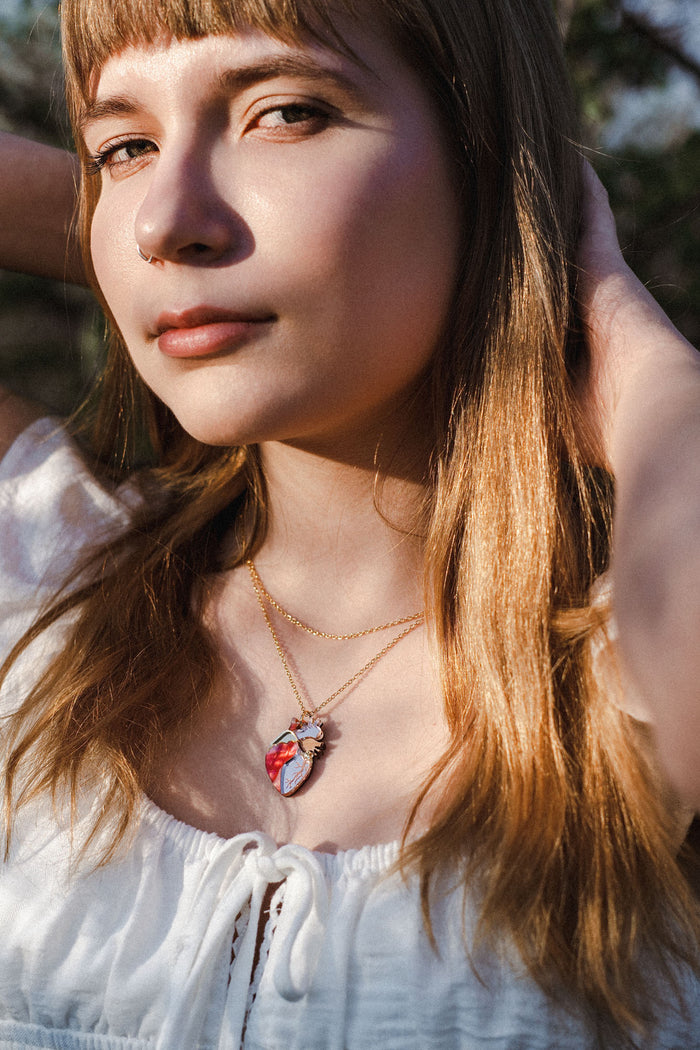 A young woman with long light brown hair wearing a white top and layered gold necklaces, including a unique anatomical heart necklace, stands outdoors with sunlight on her face and hands behind her head.