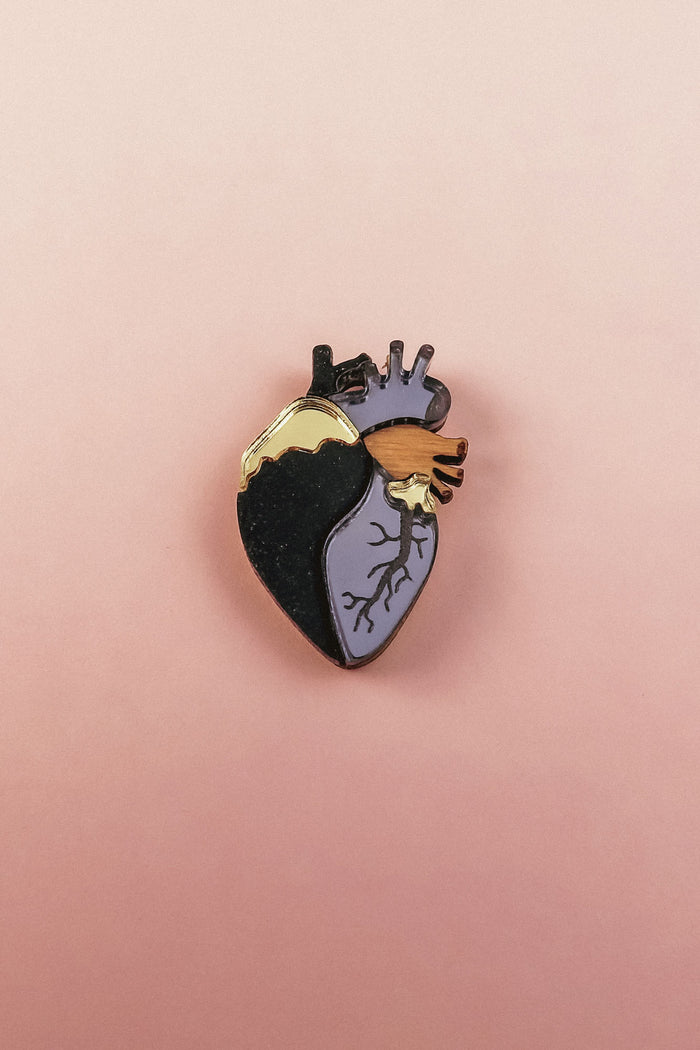 A handcrafted Anatomical Heart Pin with one side black, the other lavender, and highlighted by gold and purple accents. This statement piece rests on a pink background, perfect for adding bold flair to your collection.