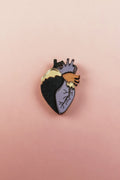 A handcrafted Anatomical Heart Pin with one side black, the other lavender, and highlighted by gold and purple accents. This statement piece rests on a pink background, perfect for adding bold flair to your collection.