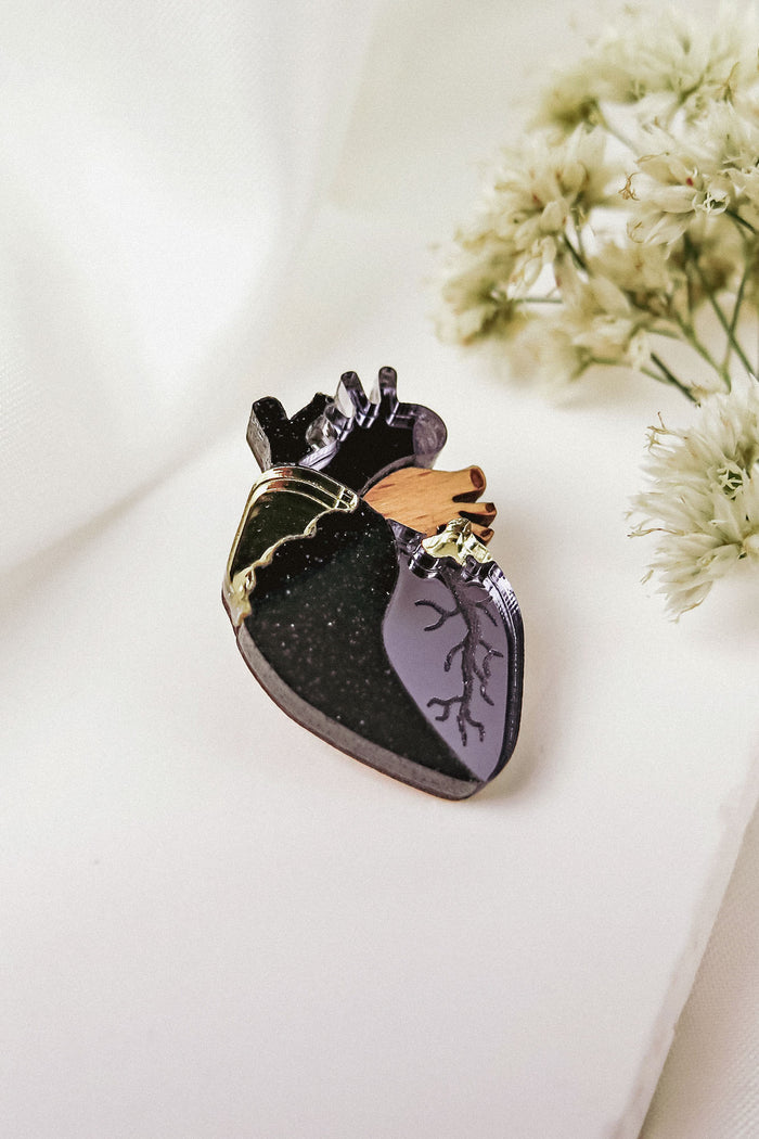 A handcrafted anatomical heart pin with black, purple, gold, and brown details rests on a white surface beside delicate white flowers—a striking statement piece for any collection.