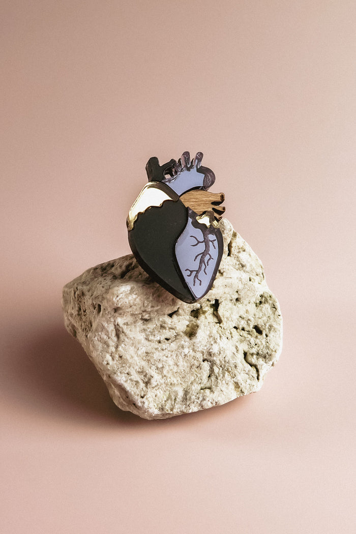 A handcrafted black and gold Anatomical Heart Pin rests on a textured light-colored rock against a plain beige background, making it a true statement piece.