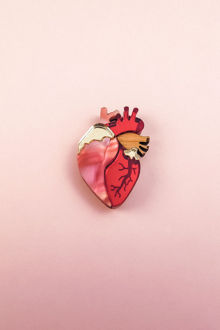 A handcrafted Anatomical Heart Pin, shaped like a human heart and featuring pink, red, and gold tones, rests on a soft pink background—perfect as a unique statement piece.