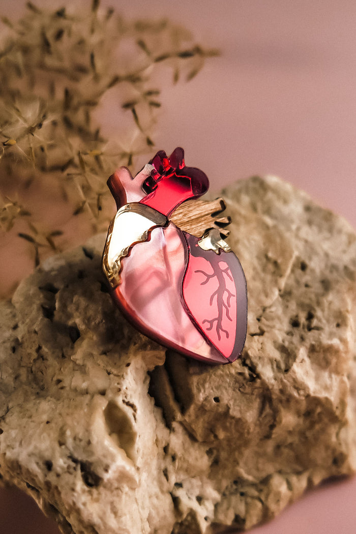 A handcrafted Anatomical Heart Pin, featuring red, pink, and gold details, rests on a textured beige rock with a blurred plant in the background—a true statement piece for any collection.