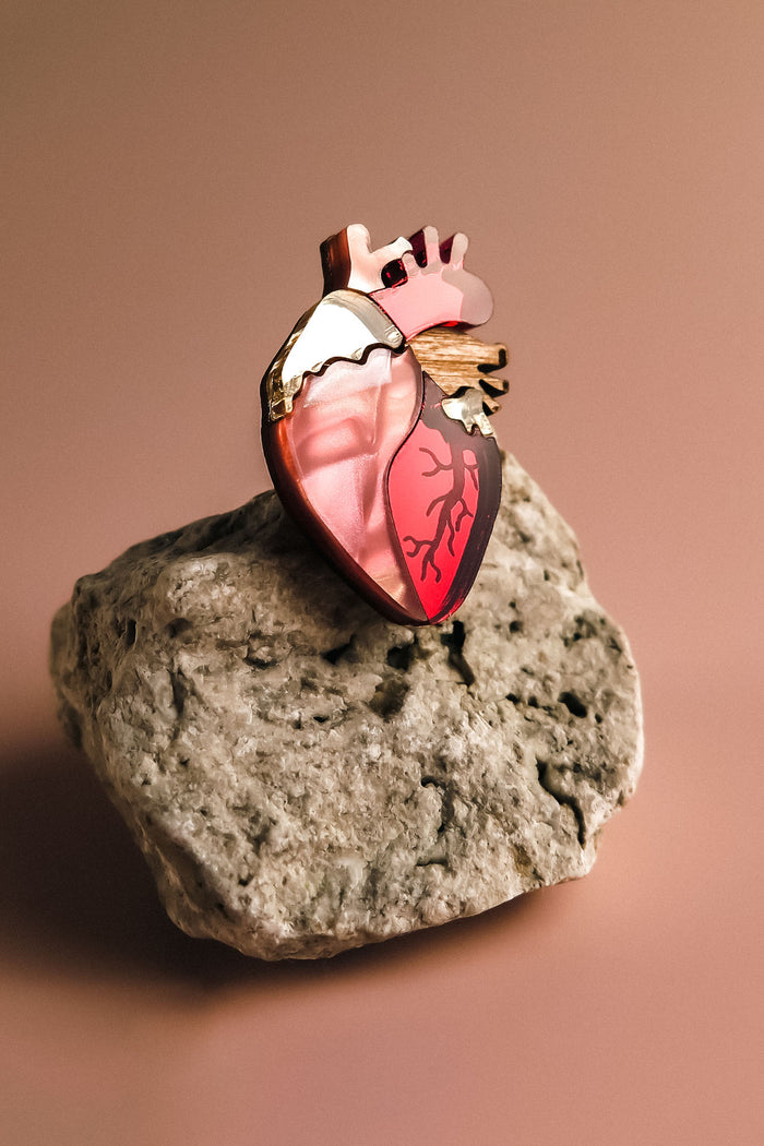 A handcrafted Anatomical Heart Pin, adorned with red and gold accents, serves as a striking statement piece atop a rough, light-colored stone against a soft brown background.
