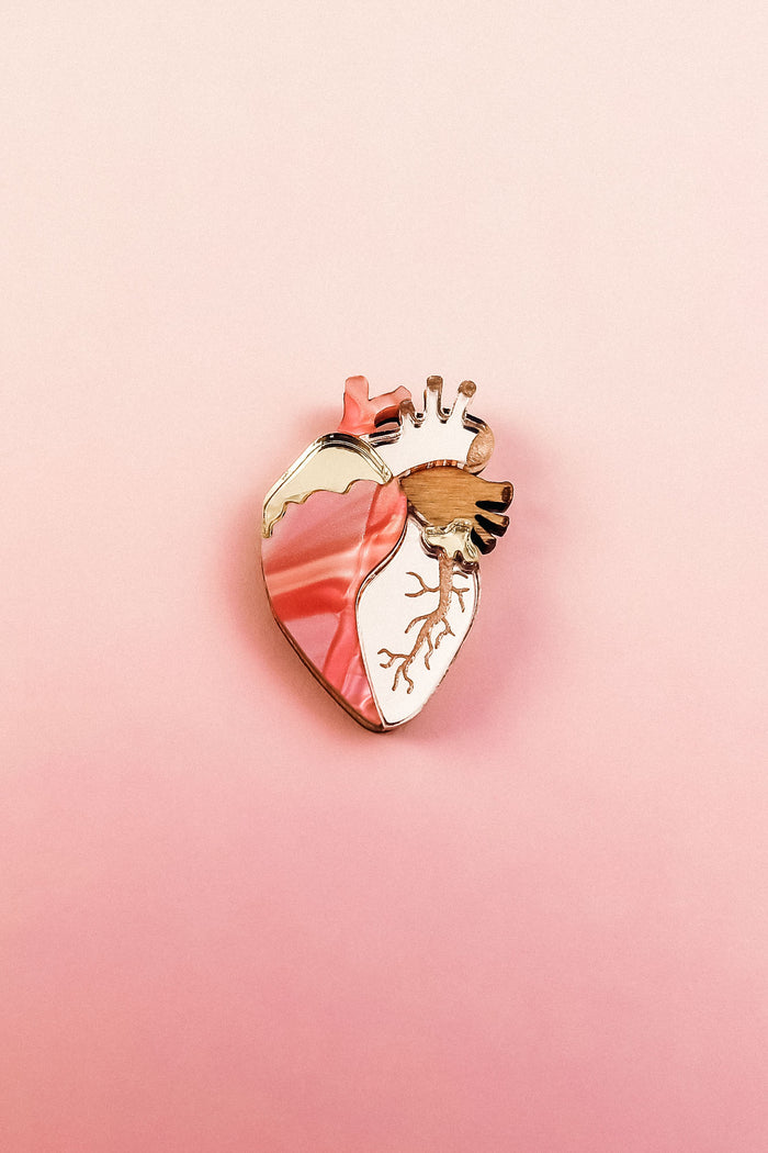 A unique, handcrafted Anatomical Heart Pin with gold accents is displayed on a pink gradient background, making it a striking decorative accessory.