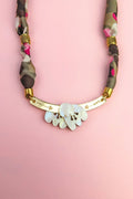 A handmade necklace with a gold curved bar, white shell-like pieces, and a multicolored fabric chain reminiscent of a silk-print scarf in green, pink, brown, and gold accents, displayed on a light pink background.