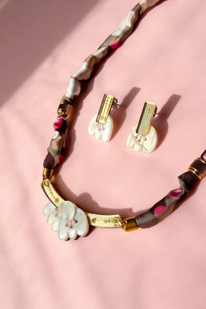 A handmade necklace and matching clip-on earrings with white, acrylic tulip-shaped pendants, gold accents, and a fabric-wrapped brown and pink chain, displayed on a soft pink background.