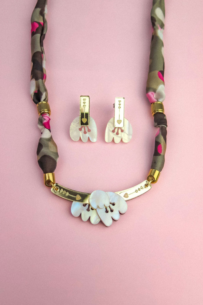 A handmade necklace with a fabric-wrapped chain and matching earrings, both featuring white, abstract, acrylic tulip-like pendants, displayed on a pink background.