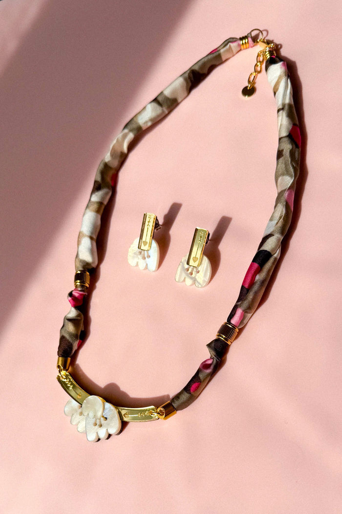 A handmade necklace and matching earrings are displayed on a pink surface. The necklace features a patterned fabric cord, gold accents, and a white floral pendant, while the earrings have white floral pieces with gold details and acrylic tulip touches.