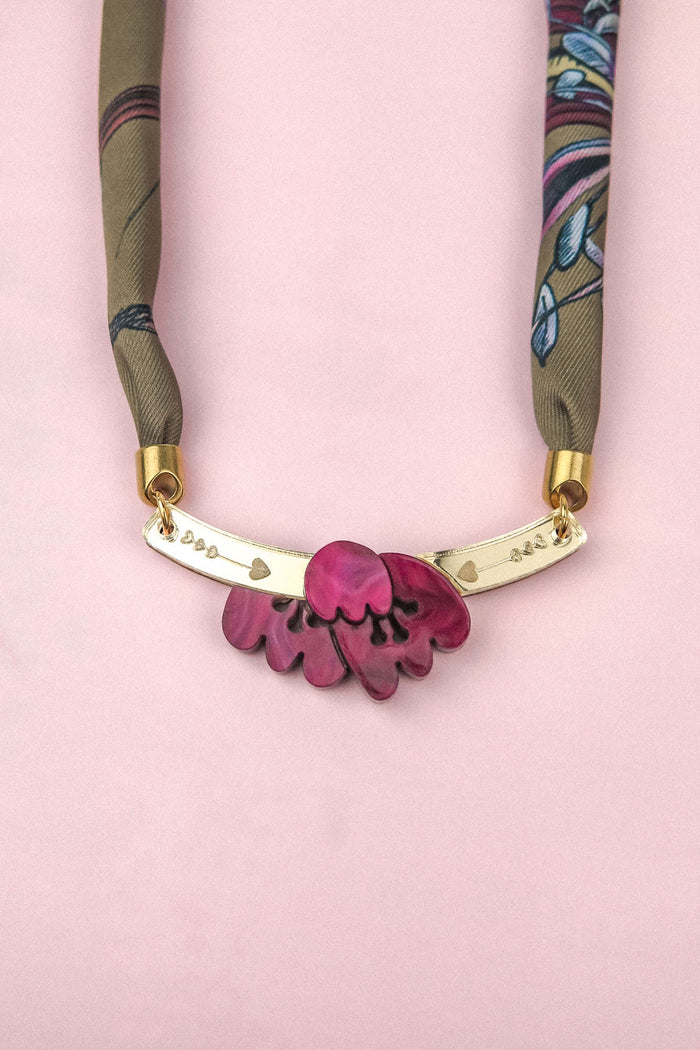 A handmade necklace featuring a pink flower pendant reminiscent of an acrylic tulip, paired with a gold bar adorned with heart and dot engravings, and suspended on a green and blue patterned silk-print scarf chain, displayed elegantly on a pale pink background.