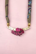 A handmade necklace featuring a pink flower pendant reminiscent of an acrylic tulip, paired with a gold bar adorned with heart and dot engravings, and suspended on a green and blue patterned silk-print scarf chain, displayed elegantly on a pale pink background.