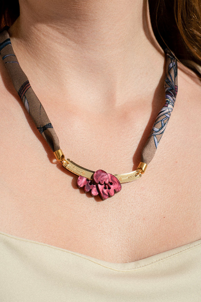 A close-up of a person wearing a handmade necklace featuring a gold curved bar, a pink floral-shaped centerpiece reminiscent of an acrylic tulip, and a patterned fabric strap around the neck. The person is dressed in a light-colored, sleeveless top.