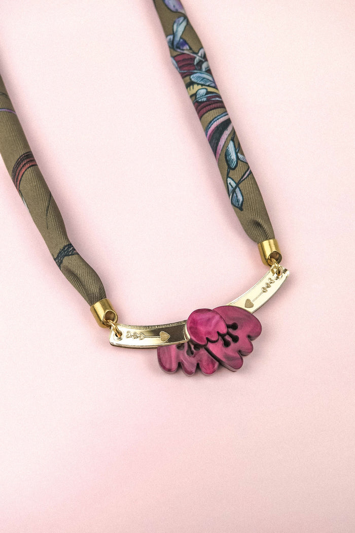 A handmade necklace featuring a fabric band reminiscent of a silk-print scarf, with gold accents and an acrylic tulip pink floral pendant, displayed on a light pink background.