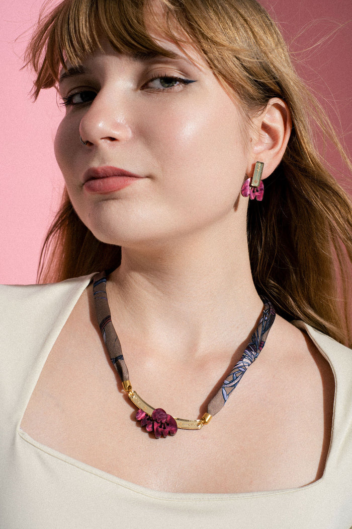 Woman with long light brown hair wearing a cream top, gold and purple earrings, and a handmade necklace with a gold bar and purple flower accents, posing against a pink background.