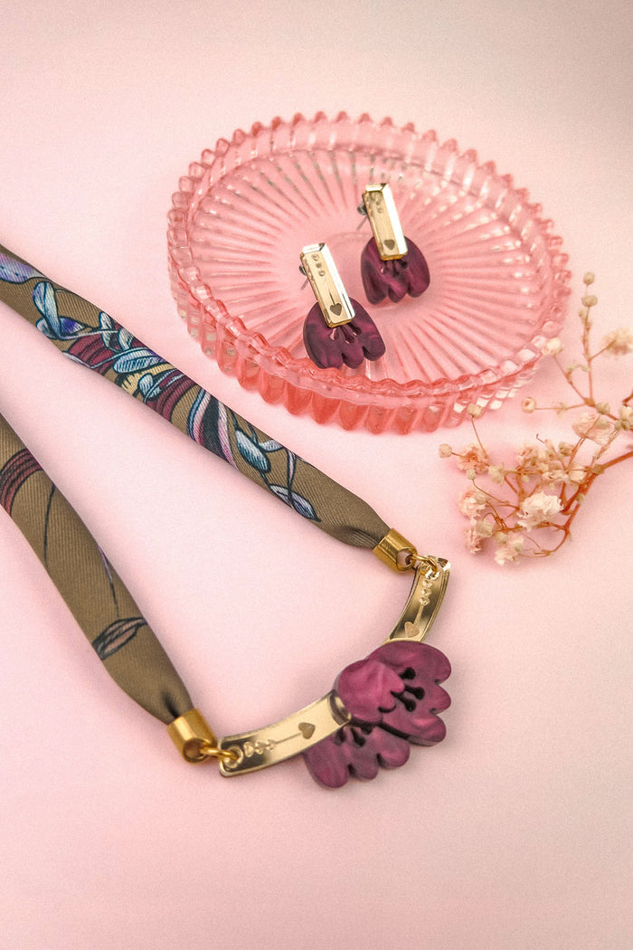 Purple flower necklace with a patterned fabric chain, reminiscent of a silk-print scarf, and matching purple clip-on earrings in a pink glass dish, with small dried flowers on a soft pink background.
