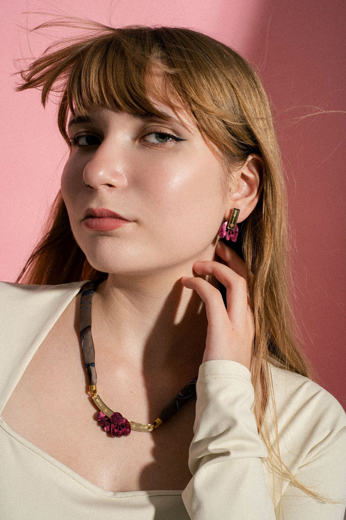 Young woman with long light brown hair and bangs poses against a pink background, wearing a cream top, a handmade necklace with matching earrings featuring purple accents, touching her neck with one hand and looking confidently at the camera.