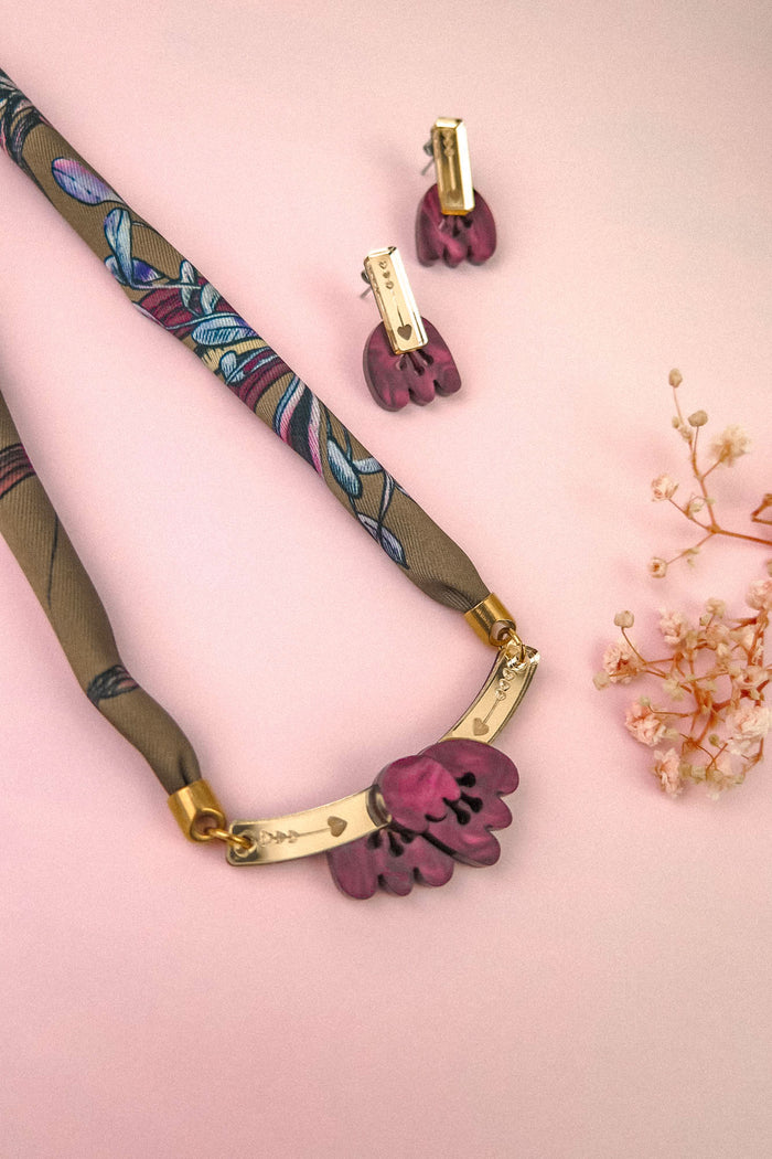 A handmade necklace with floral details and matching earrings rests on a pink surface, beside small white dried flowers.
