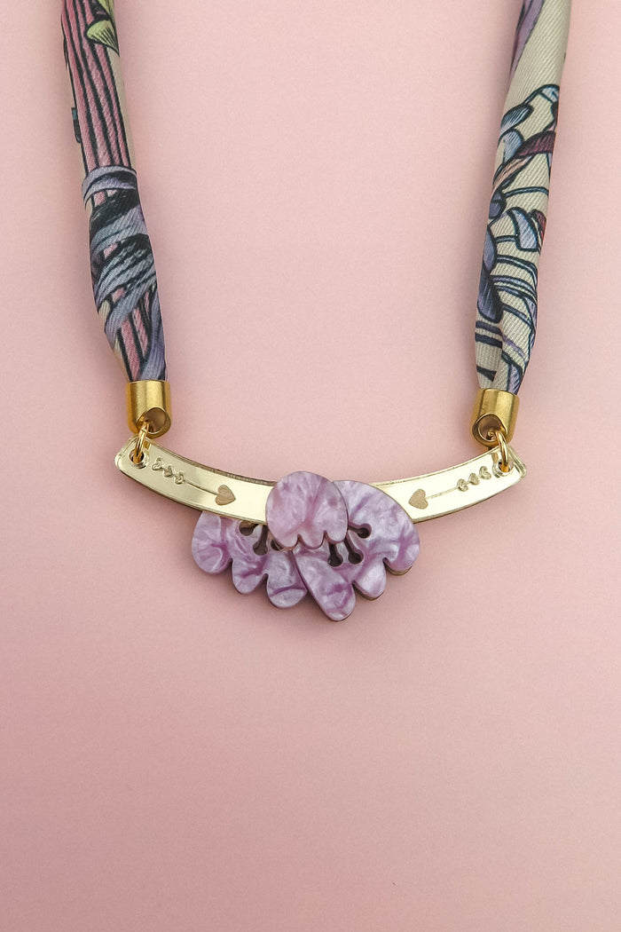 A handmade necklace featuring a pink and purple patterned fabric strap, a gold curved bar with small heart cutouts, and a cluster of marbled lavender decorative stones, displayed on a light pink background.