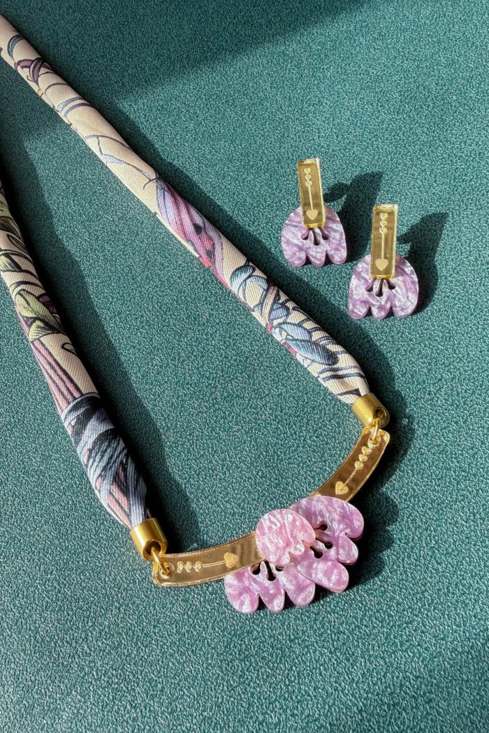 A handmade necklace with a patterned fabric cord, gold metal bar, and pink acrylic tulip pendants is displayed next to matching pink floral earrings on a textured teal surface.