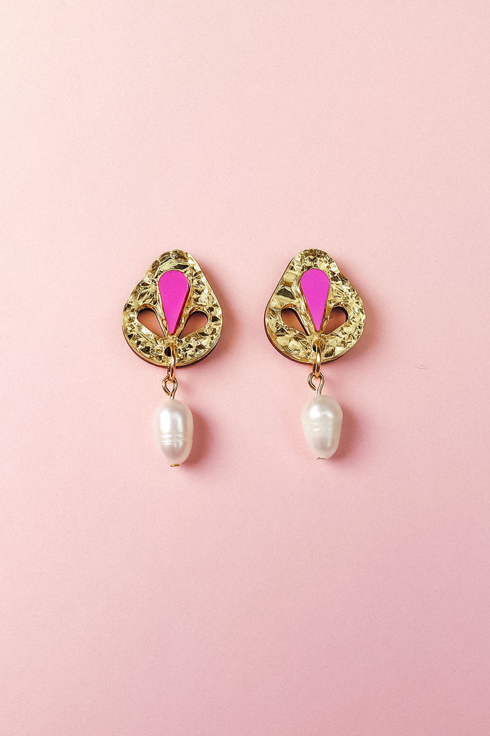 A pair of handcrafted gold drop earrings with intricate designs, featuring a pink enamel detail at the top and a white pearl dangling at the bottom, displayed on a light pink background.