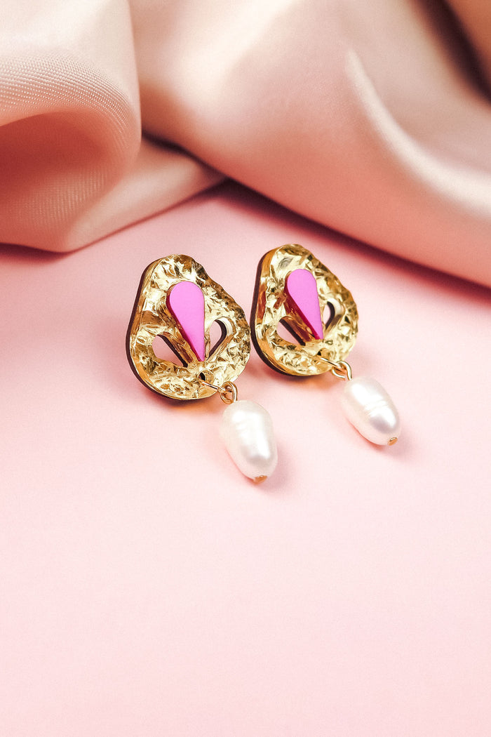 Handcrafted gold drop earrings with textured, abstract shapes, featuring pink and magenta accents and a dangling white pearl—perfect for minimalist fashion—displayed on a soft pink surface with silky pink fabric in the background.