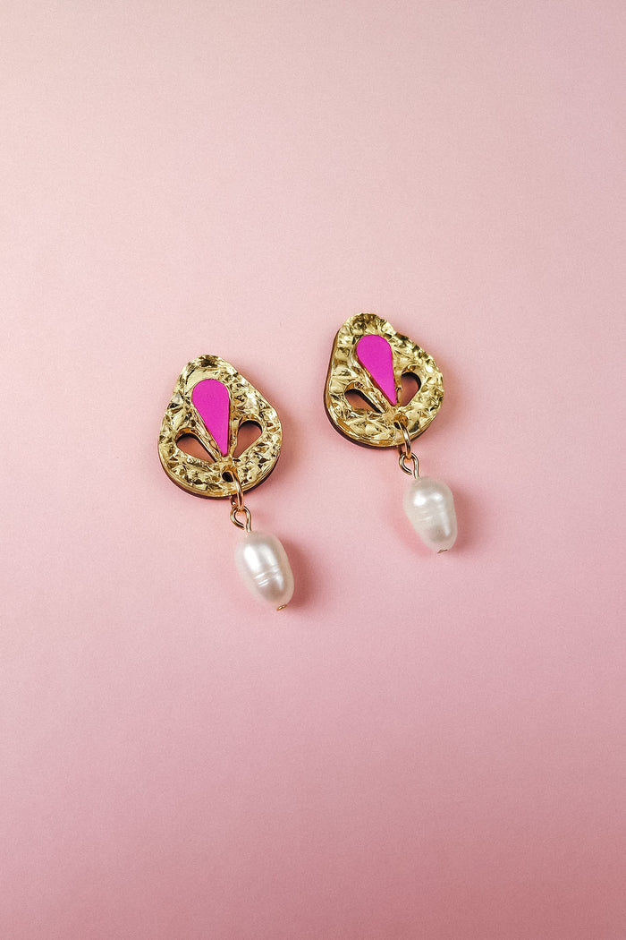 A pair of handcrafted gold-toned drop earrings with pink accents and a dangling white pearl, displayed on a soft pink background.
