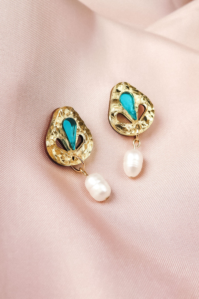 A pair of handcrafted gold earrings with blue enamel accents and dangling white pearls, displayed on light pink satin fabric—perfect for elevating your minimalist fashion style.