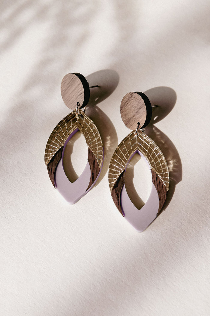 A pair of stylish, handcrafted drop earrings featuring layered geometric shapes in wood, gold, and premium acrylic glass in lavender, casting soft shadows on a light surface.