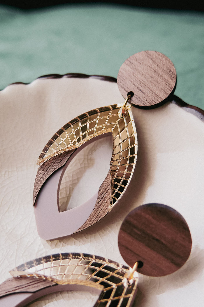 Close-up of handcrafted earrings with circular wooden studs and geometric drop pendants featuring gold lattice, wood accents, and muted purple elements, displayed on a cream-colored ceramic dish. Made with premium acrylic glass details.