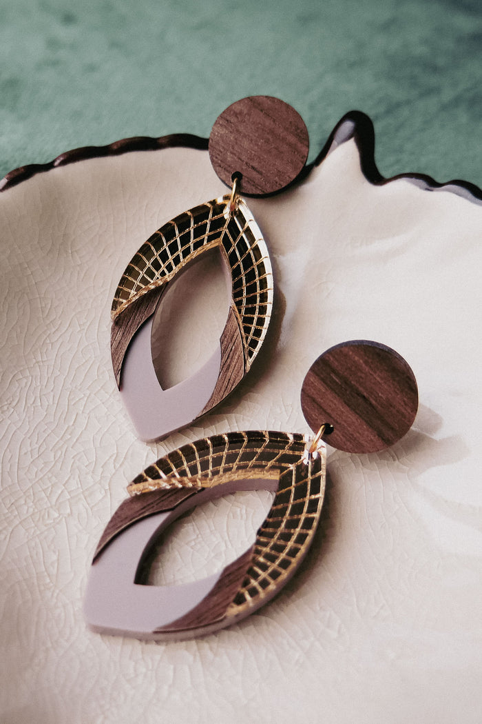A pair of stylish handcrafted earrings with wooden circular studs and layered geometric dangles featuring gold, brown wood, and light beige accents in premium acrylic glass, displayed on a textured cream-colored dish.