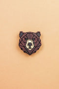 A handcrafted wooden brooch in a geometric style, shaped like a bear’s head with dark brown and light beige sections, placed on a plain light orange background.