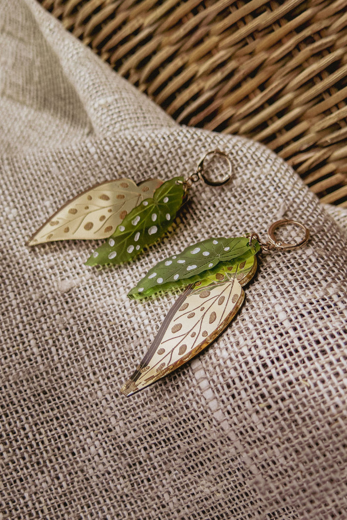 A pair of Begonia Leaves Earrings, perfect for plant lovers, shaped like leaves with gold and green hues and white dots, displayed on beige textured fabric next to a woven basket.