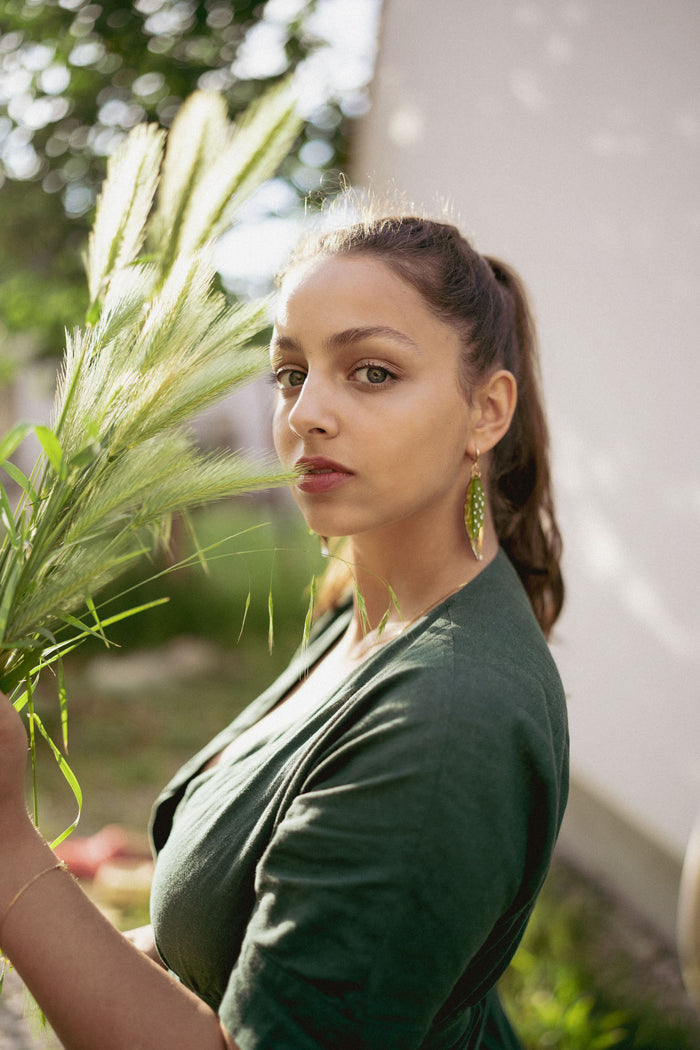 A woman with long brown hair in a ponytail, wearing a dark green top and unique jewelry—Begonia Leaves Earrings—stands outdoors in sunlight holding tall grass, looking directly at the camera. The softly blurred background is perfect for plant lovers.
