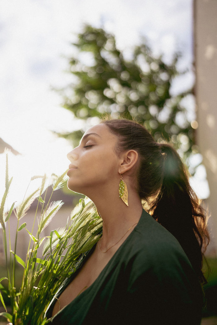 A woman with long dark hair in a ponytail stands outdoors, eyes closed, holding green plants. Sunlight softly illuminates her face as she wears unique jewelry—a large Begonia Leaves Earring. A blurred tree appears in the background, perfect for plant lovers.