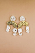 A pair of abstract dangle earrings with textured gold curved pieces, irregular white shapes, and small embedded red and green rhinestones, displayed on a beige background.