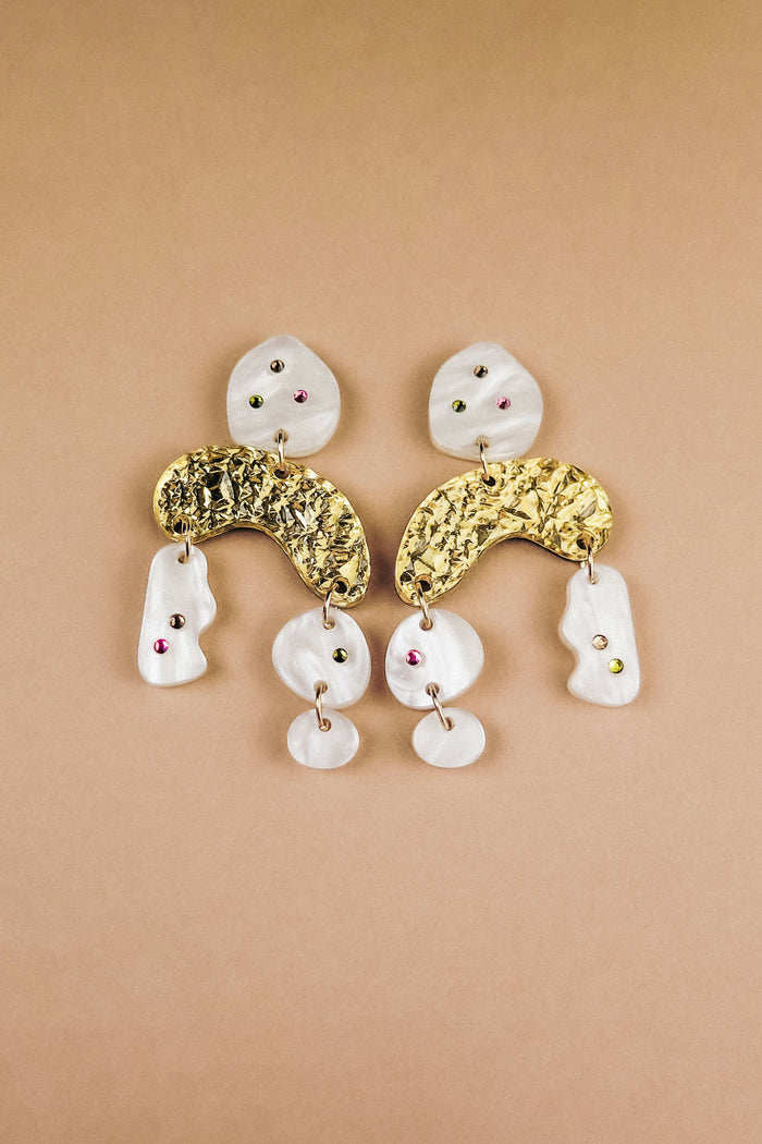 A pair of abstract dangle earrings with textured gold curved pieces, irregular white shapes, and small embedded red and green rhinestones, displayed on a beige background.