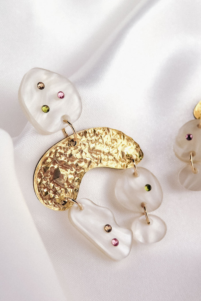 A close-up of a boho chic dangle earring featuring textured gold metal and irregularly shaped white resin pieces adorned with small pink and green rhinestones, set against a soft white satin background.