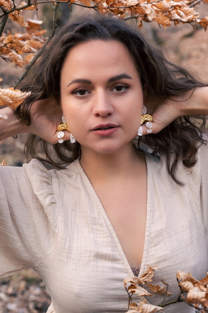 A woman with wavy brown hair poses outdoors among dry autumn leaves, wearing a light beige dress and boho chic dangle earrings adorned with rhinestones, her hands touching her hair and an intense, confident expression on her face.